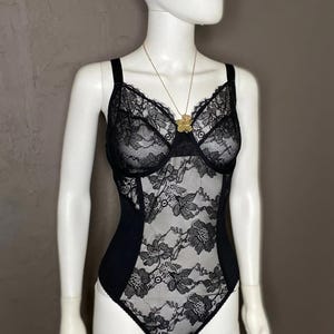 Könnte beinhalten: Schwarzer Spitzenbody mit floralen Mustern, präsentiert auf einer Schaufensterpuppe. Der Body hat verstellbare Träger und einen Sweetheart-Ausschnitt. Eine goldfarbene Halskette mit einem Blumenanhänger ist ebenfalls zu sehen.
