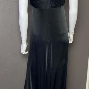 K&ouml;nnte beinhalten: Ein bodenlanges, schwarzes Kleid an einer Schaufensterpuppe. Das Kleid hat einen V-Ausschnitt und eine geraffte Taillenpartie. Die Schaufensterpuppe ist wei&szlig;, und eine goldfarbene Halskette ist sichtbar. Der Hintergrund ist eine graue Wand.