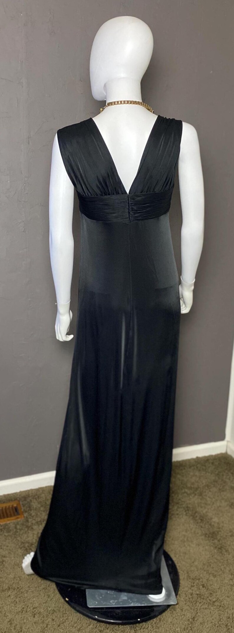 K&ouml;nnte beinhalten: Ein bodenlanges, schwarzes Kleid an einer Schaufensterpuppe. Das Kleid hat einen V-Ausschnitt und eine geraffte Taillenpartie. Die Schaufensterpuppe ist wei&szlig;, und eine goldfarbene Halskette ist sichtbar. Der Hintergrund ist eine graue Wand.