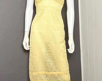 May include: A knee-length yellow slip dress featuring an all-over floral lace overlay, a V-neckline with scalloped lace trim, and wide shoulder straps. The garment includes a horizontal lace band detail near the hemline and a scalloped lace edge.