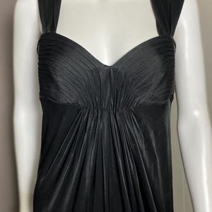 K&ouml;nnte beinhalten: Ein schwarzes Abendkleid mit herzf&ouml;rmigem Ausschnitt und plissiertem Oberteil. Das Kleid hat eine figurbetonte Silhouette und einen flie&szlig;enden Rock. Eine goldfarbene Halskette mit dunklen Akzenten schm&uuml;ckt den Ausschnitt. Das Kleid wird auf einer Schaufensterpuppe pr&auml;sentiert.