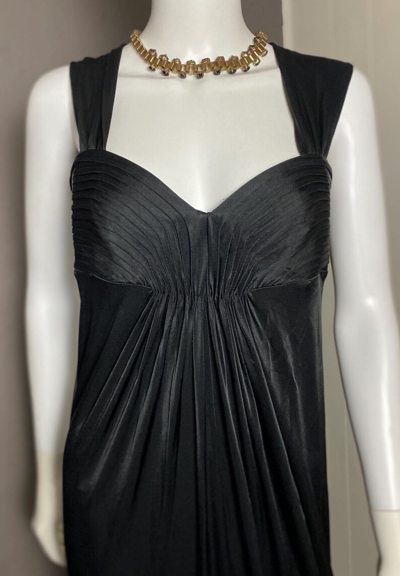 K&ouml;nnte beinhalten: Ein schwarzes Abendkleid mit herzf&ouml;rmigem Ausschnitt und plissiertem Oberteil. Das Kleid hat eine figurbetonte Silhouette und einen flie&szlig;enden Rock. Eine goldfarbene Halskette mit dunklen Akzenten schm&uuml;ckt den Ausschnitt. Das Kleid wird auf einer Schaufensterpuppe pr&auml;sentiert.