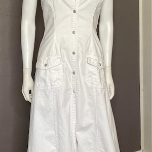 Op de afbeelding: Witte denim jumpsuit met een halterhalslijn, knoopsluiting aan de voorkant en twee voorzakken. De jumpsuit wordt op een mannequin gepresenteerd. Het kledingstuk heeft een getailleerd lijfje en een wijd model.