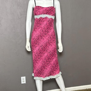 May include: A knee-length slip dress featuring a vibrant pink and black snakeskin pattern. The garment includes thin black spaghetti straps, a straight neckline, and white lace trim detailing along the bodice and the bottom hemline.