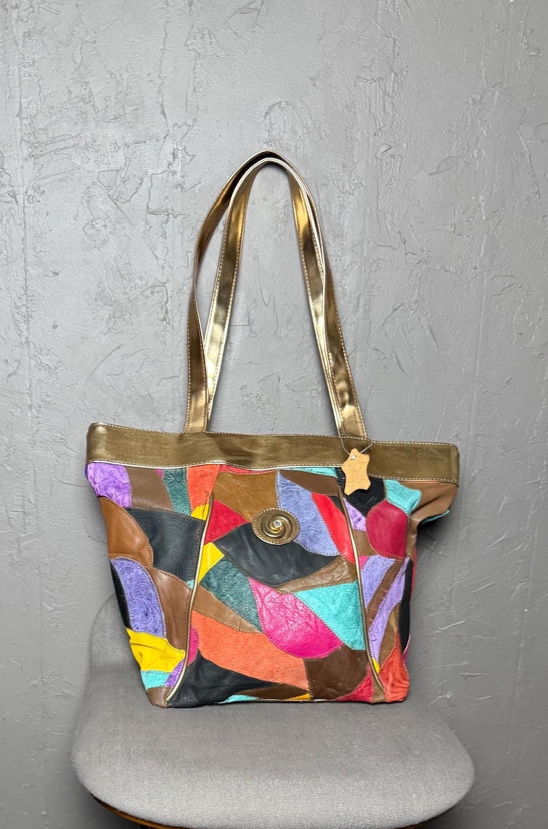 Peut inclure: Un sac fourre-tout en cuir patchwork multicolore avec des anses dor&eacute;es. Le sac pr&eacute;sente des pi&egrave;ces de cuir dans les tons de violet, rouge, orange, jaune et noir. Un fermoir d&eacute;coratif en m&eacute;tal est sur le devant.
