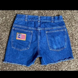 以下が含まれることがあります： 後ろのポケットに「76」の文字が入った小さなアメリカ国旗のパッチが付いた、裾がフリンジになった青いデニムのショートパンツです。