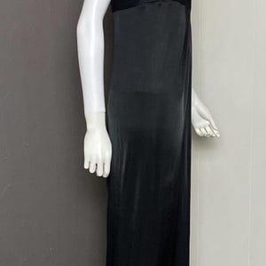K&ouml;nnte beinhalten: Schwarzes Abendkleid an einer Schaufensterpuppe. Das Kleid hat ein plissiertes Oberteil und einen flie&szlig;enden, bodenlangen Rock. Eine goldfarbene Halskette schm&uuml;ckt den Hals der Schaufensterpuppe. Das Kleid wird vor einem neutralen Hintergrund pr&auml;sentiert.