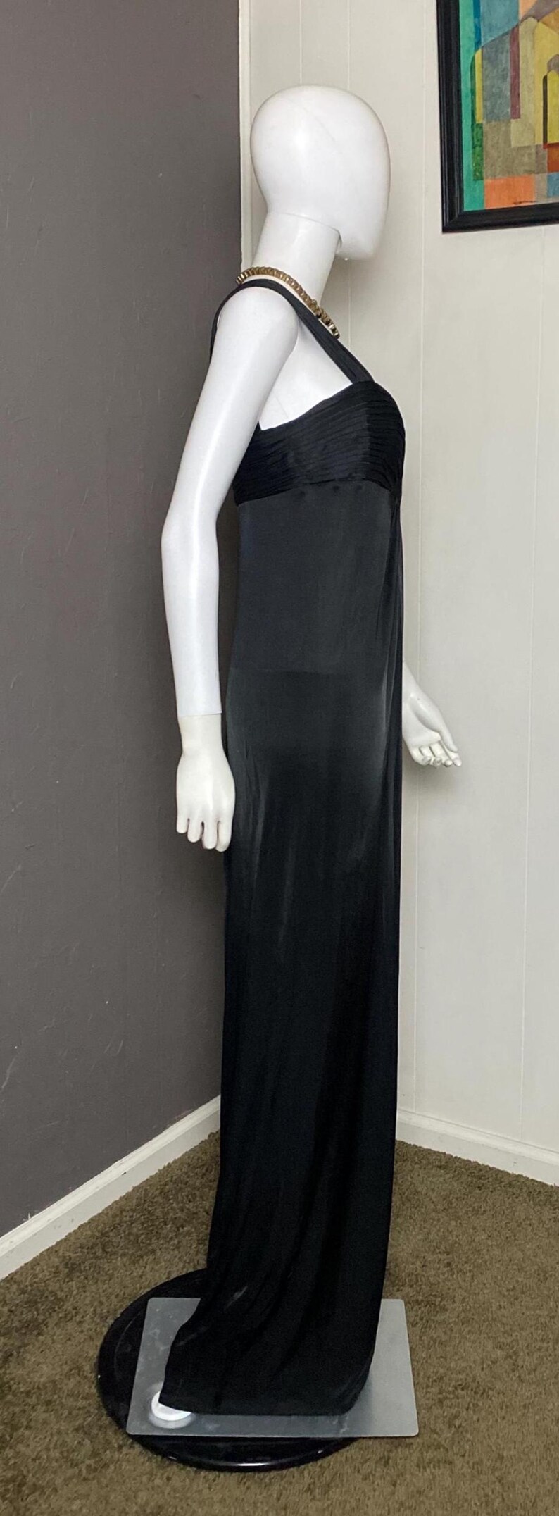 K&ouml;nnte beinhalten: Schwarzes Abendkleid an einer Schaufensterpuppe. Das Kleid hat ein plissiertes Oberteil und einen flie&szlig;enden, bodenlangen Rock. Eine goldfarbene Halskette schm&uuml;ckt den Hals der Schaufensterpuppe. Das Kleid wird vor einem neutralen Hintergrund pr&auml;sentiert.