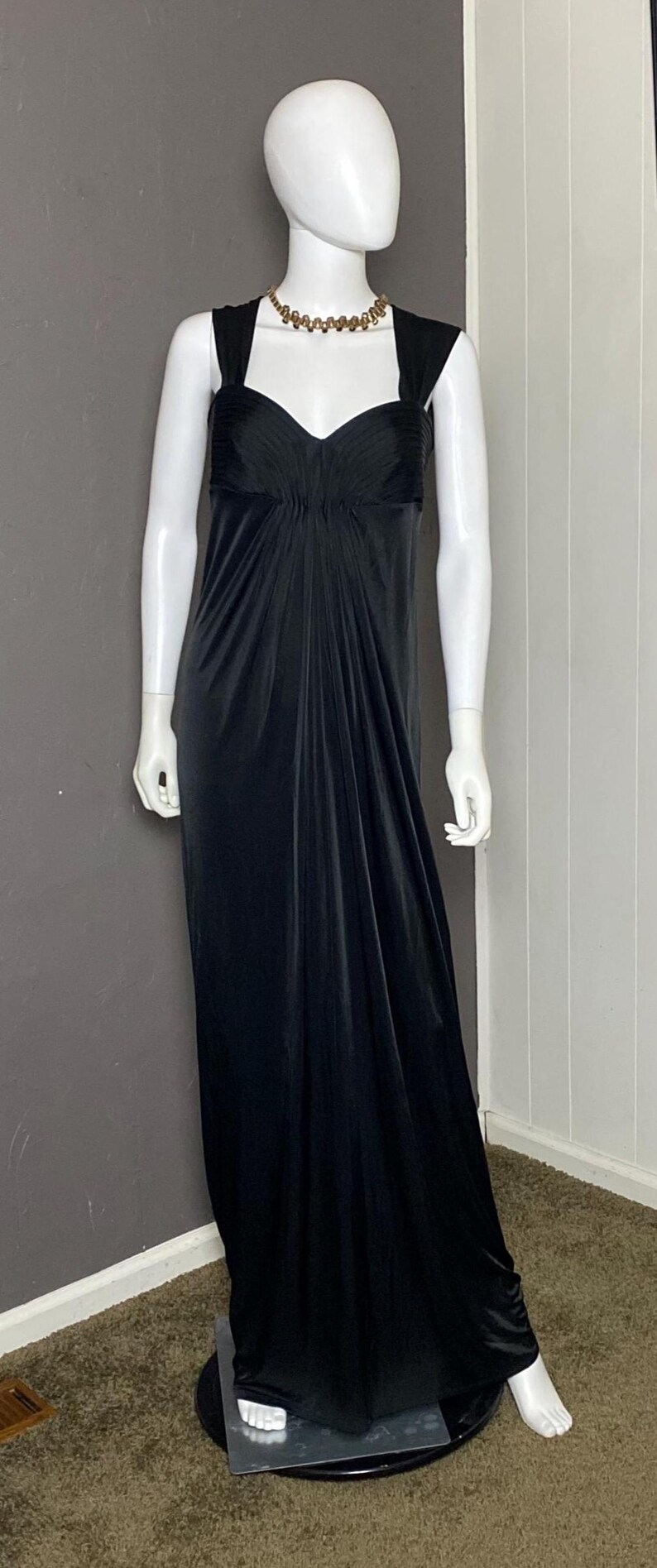 K&ouml;nnte beinhalten: Ein schwarzes Abendkleid auf einer Schaufensterpuppe. Das Kleid hat einen herzf&ouml;rmigen Ausschnitt, ein eng anliegendes Oberteil und einen flie&szlig;enden Rock. Eine goldfarbene Halskette schm&uuml;ckt den Hals der Schaufensterpuppe. Das bodenlange Kleid ist aus weichem, drapierendem Stoff gefertigt.