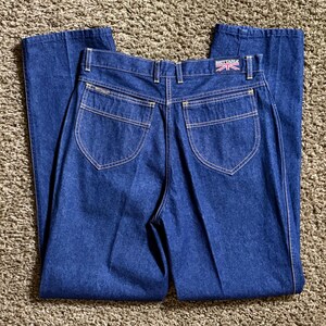 Peut inclure: Jean bleu foncé avec une taille haute et une jambe évasée. Les poches arrière ont un design arrondi avec des coutures blanches. Le nom de la marque "BRITTANIA" est visible sur la ceinture arrière.