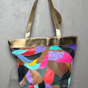 Peut inclure: Un sac fourre-tout patchwork multicolore avec des poign&eacute;es et des bordures dor&eacute;es. Le sac pr&eacute;sente des pi&egrave;ces de cuir rouges, violettes, roses, marron et noires. Le sac est con&ccedil;u pour transporter des objets.