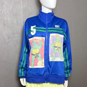 May include: A blue zip-up track jacket featuring green stripes on the sleeves, a white number 5 patch on the chest, and two rectangular graphic prints of Teenage Mutant Ninja Turtles characters. The jacket has a high collar and a small logo patch.