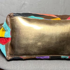 Peut inclure: Un sac &agrave; main patchwork color&eacute; avec une base dor&eacute;e. Le sac pr&eacute;sente une vari&eacute;t&eacute; de couleurs, dont le rouge, le bleu, le jaune et le marron. Il a une poign&eacute;e et une petite &eacute;tiquette en cuir attach&eacute;e. Le sac est pos&eacute; sur une surface grise.