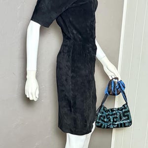May include: A black suede dress with short sleeves and a fitted silhouette. The dress is displayed on a mannequin. A small, patterned handbag with blue accents is held by the mannequin.