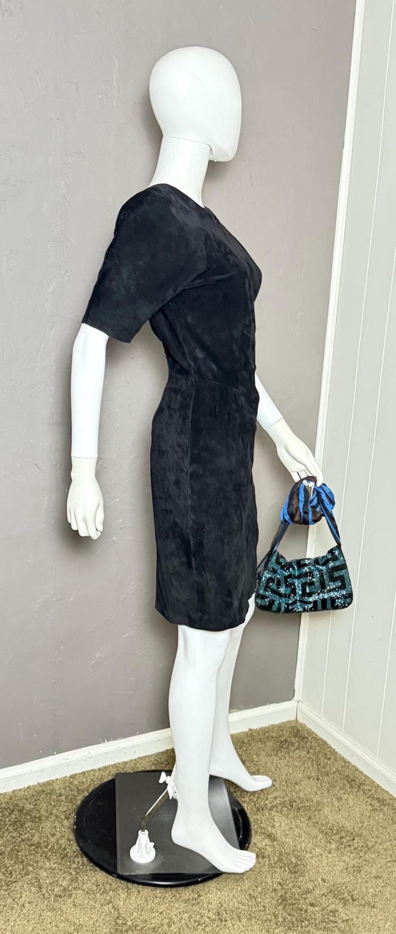 May include: A black suede dress with short sleeves and a fitted silhouette. The dress is displayed on a mannequin. A small, patterned handbag with blue accents is held by the mannequin.