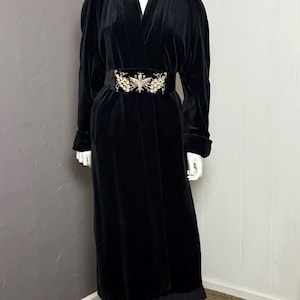 May include: A long, black velvet robe with a decorative belt featuring an ornate gold and silver design. The robe is displayed on a mannequin. The hemline of the robe is just above the ankles.