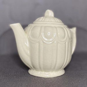 May include: A white ceramic teapot with a textured, scalloped design. The teapot has a rounded body and a lid with a knob on top.