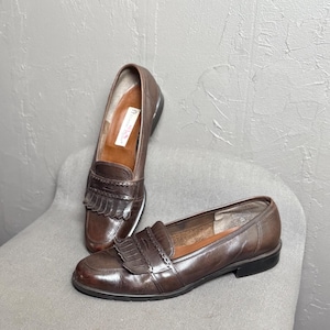 May include: A pair of brown leather loafers. These shoes have a classic design with a rounded toe, decorative fringe, and a low heel. The loafers are displayed on a grey chair against a textured grey background.