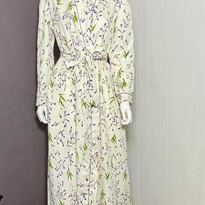 Puede incluir: Bata color crema con un estampado floral con detalles en morado y verde. La bata tiene un cinturón a juego, mangas largas y un ribete morado oscuro en contraste. La bata se exhibe en un maniquí.