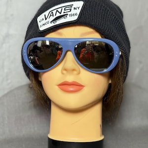 Può includere: Una testa di manichino con un berretto di maglia nero con una toppa Vans e occhiali da sole azzurri. Gli occhiali da sole hanno una montatura arrotondata e lenti scure. La toppa Vans recita "CA Vans NY Since 1966".