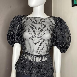 May include: A black sheer blouse featuring puff sleeves and a boat neckline. The garment is decorated with intricate, wavy black beaded patterns across the bodice and sleeves, with a gathered peplum waist and a solid black skirt underneath.