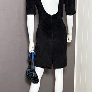 May include: A black dress with a low back and short sleeves is displayed on a mannequin. The dress has a slit in the back and is paired with a small, sequined purse with blue accents.