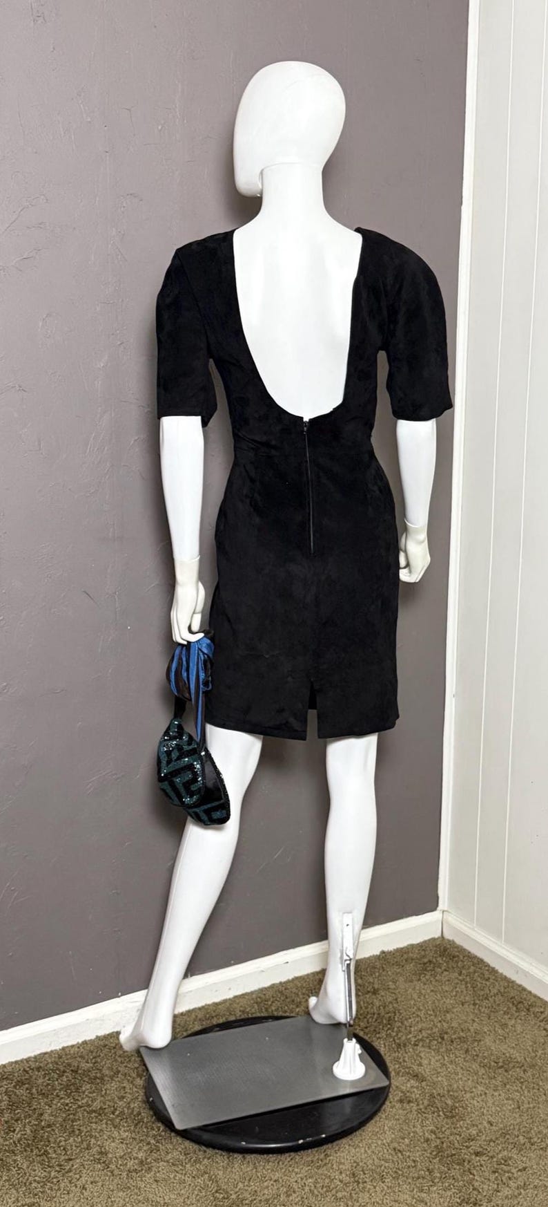 May include: A black dress with a low back and short sleeves is displayed on a mannequin. The dress has a slit in the back and is paired with a small, sequined purse with blue accents.
