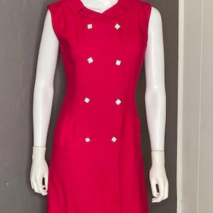 May include: A sleeveless pink dress with a double-breasted button closure and white buttons. The dress has a collar and a fitted silhouette.