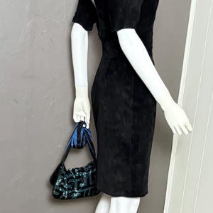May include: A black, short-sleeved dress with a knee-length hem is displayed on a mannequin. The dress is paired with a small, sequined handbag with blue accents. The mannequin is on a black base, set against a gray wall.