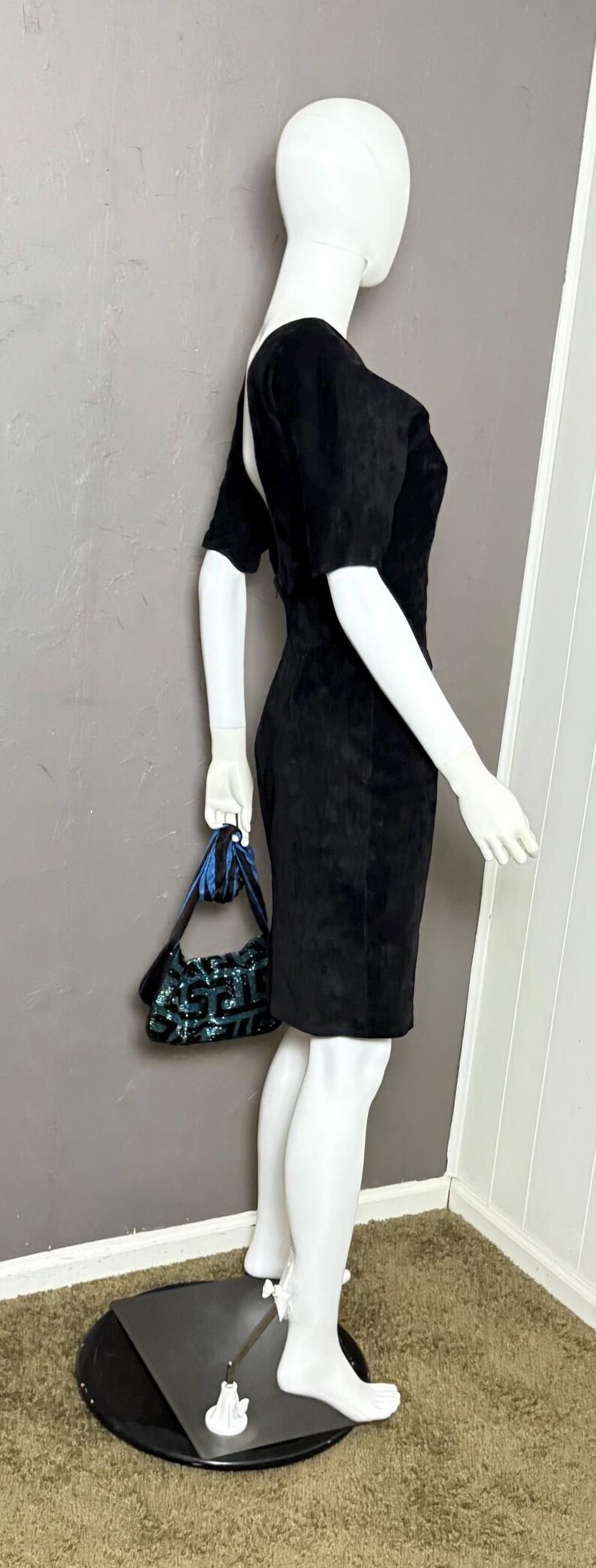 May include: A black, short-sleeved dress with a knee-length hem is displayed on a mannequin. The dress is paired with a small, sequined handbag with blue accents. The mannequin is on a black base, set against a gray wall.
