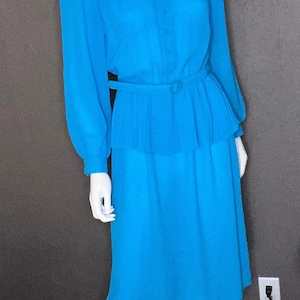 May include: A bright turquoise dress on a mannequin. The dress features long sleeves, a button-down bodice, and a belted waist. The skirt falls to the knees. The dress is a solid color and appears to be made of a lightweight fabric.