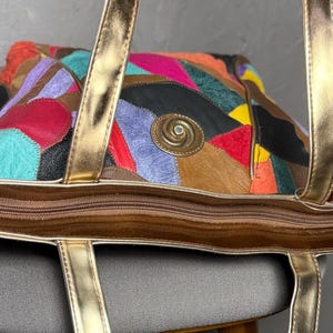 Peut inclure: Sac fourre-tout patchwork color&eacute; avec des anses dor&eacute;es et une fermeture &eacute;clair. Le sac pr&eacute;sente divers patchs en cuir dans les tons marron, rouge, bleu et violet. Un embl&egrave;me circulaire dor&eacute; est centr&eacute; sur le devant.