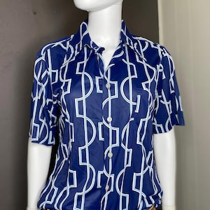 Puede incluir: Una camisa azul de manga corta con botones y un patrón geométrico blanco. La camisa tiene un bolsillo en el lado izquierdo. El maniquí lleva un cinturón marrón con una hebilla grande de metal y pantalones vaqueros azules.