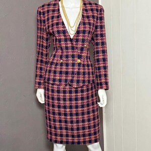 May include: A two-piece skirt suit featuring a navy blue, magenta, and orange plaid pattern. The jacket has long sleeves, a V-neckline, and four gold-toned buttons in a double-breasted arrangement. The matching skirt is knee-length and straight-cut.