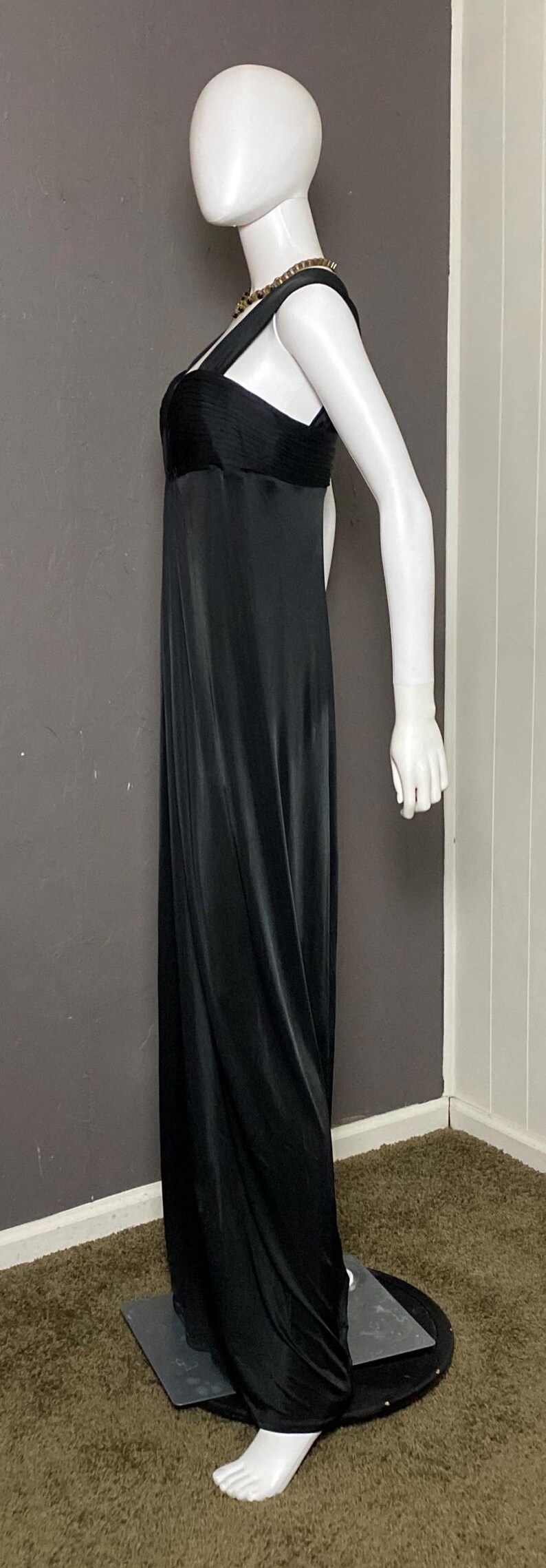 K&ouml;nnte beinhalten: Ein bodenlanges schwarzes Kleid auf einer Schaufensterpuppe. Das Kleid hat ein plissiertes Oberteil und Spaghettitr&auml;ger. Eine goldfarbene Halskette schm&uuml;ckt den Hals der Schaufensterpuppe. Das Kleid ist aus flie&szlig;endem, seidigem Material.