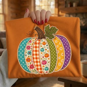 May include: A burnt orange fabric item featuring a large, central pumpkin applique. The pumpkin is composed of vertical segments with diverse patterns, including floral prints, orange gingham, polka dots, and small pumpkin motifs in various colours.