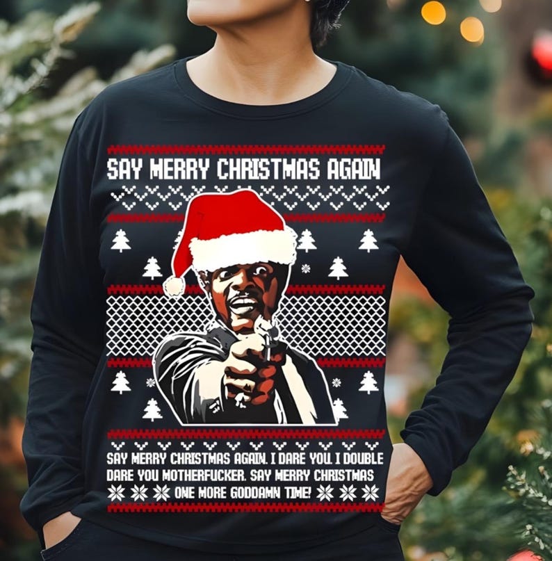 May include: Black long-sleeve shirt with a Christmas sweater design. The shirt features a graphic of a man wearing a Santa hat, holding a gun, and the text "SAY MERRY CHRISTMAS AGAIN." The design includes Christmas trees and snowflakes.