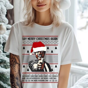 May include: White t-shirt with a humorous Christmas design. The graphic features a man wearing a Santa hat, holding a gun, and the text "SAY MERRY CHRISTMAS AGAIN." The design includes Christmas trees and a repeating text pattern.