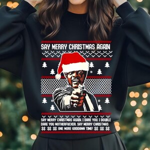 May include: Black sweatshirt with a Christmas-themed design. The design features a man wearing a Santa hat and holding a gun, with the text "SAY MERRY CHRISTMAS AGAIN." Additional text includes "I dare you...Say Merry Christmas...One more goddamn time!"