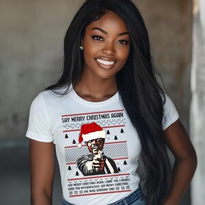 May include: White t-shirt with a graphic of a man wearing a Santa hat and pointing a gun. The text "SAY MERRY CHRISTMAS AGAIN" is above the graphic, with additional text below. The shirt has a festive, holiday theme.