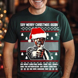 May include: Dark green t-shirt with a Christmas sweater design. The shirt features a graphic of a man wearing a Santa hat and pointing a gun. Text on the shirt reads "SAY MERRY CHRISTMAS AGAIN" and other phrases.