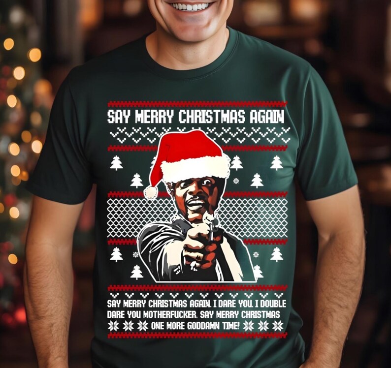 May include: Dark green t-shirt with a Christmas sweater design. The shirt features a graphic of a man wearing a Santa hat and pointing a gun. Text on the shirt reads "SAY MERRY CHRISTMAS AGAIN" and other phrases.