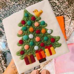 Peut inclure: Tissu crème avec une broderie représentant un sapin de Noël. L'arbre est vert avec des boules colorées et un oiseau jaune. Des cadeaux sont au pied de l'arbre, avec des boîtes rouges, oranges et jaunes.