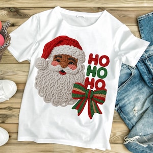 May include: White t-shirt featuring a detailed Santa Claus design with a red hat, a white beard, and the text "HO HO HO" in red and green. A festive red and green bow completes the Christmas theme.