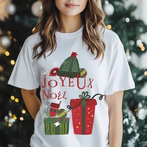 May include: White t-shirt with a festive Christmas design. The graphic includes a green knit hat, snowflakes, and the words "JOYEUX Noël" in red. Below are illustrated gift boxes and Christmas ornaments, creating a holiday theme.