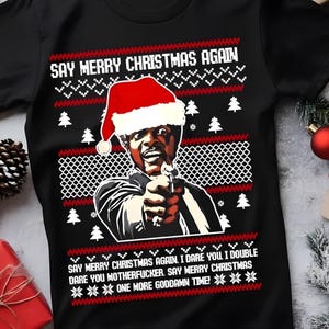 May include: Black t-shirt with a Christmas sweater design. The graphic features a man wearing a Santa hat, pointing a gun, and the text "SAY MERRY CHRISTMAS AGAIN." Additional text includes "I dare you..." and "One more goddamn time!"