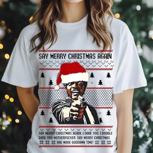 May include: White t-shirt with a Christmas-themed graphic. The design features a man wearing a Santa hat, holding a gun, and the text "SAY MERRY CHRISTMAS AGAIN." Additional text at the bottom reads "ONE MORE GODDAMN TIME!"