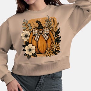 Peut inclure: Sweat-shirt beige avec un motif sur le thème de l'automne. Le motif comprend une citrouille orange avec un nœud à carreaux, entourée de fleurs et de feuillage. Le sweat-shirt a un col rond et des manches longues.