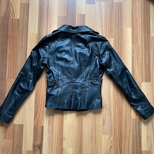 K&ouml;nnte beinhalten: Schwarze Lederjacke mit Reverskragen, langen &Auml;rmeln und aufgesetzten Taschen. Die R&uuml;ckseite der Jacke hat ein ma&szlig;geschneidertes Design mit vertikalen N&auml;hten und einem kleinen Schlitz am Saum. Die Jacke wird auf einer Holzoberfl&auml;che pr&auml;sentiert.