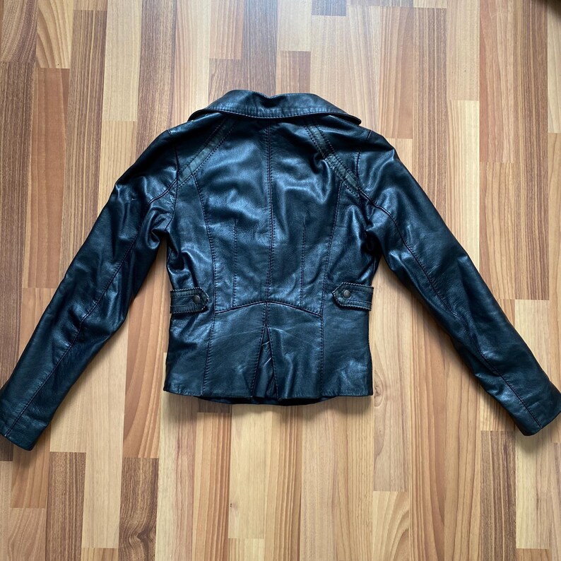 K&ouml;nnte beinhalten: Schwarze Lederjacke mit Reverskragen, langen &Auml;rmeln und aufgesetzten Taschen. Die R&uuml;ckseite der Jacke hat ein ma&szlig;geschneidertes Design mit vertikalen N&auml;hten und einem kleinen Schlitz am Saum. Die Jacke wird auf einer Holzoberfl&auml;che pr&auml;sentiert.